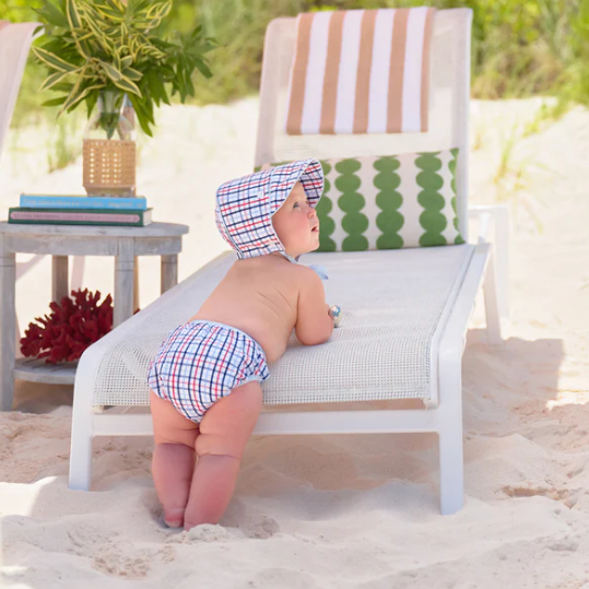 Baby in swimwear and hat sitting on a beach chair under a striped umbrella.